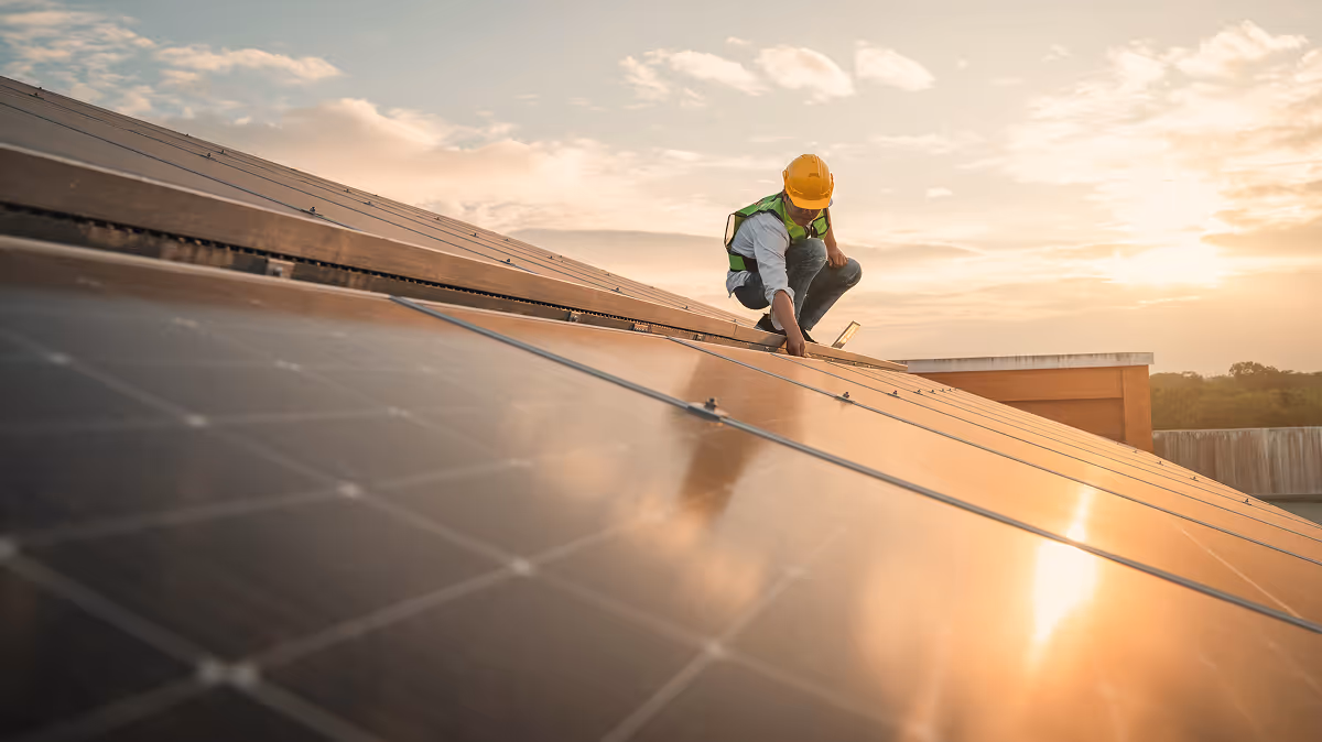 Solar technician in a yellow safety helmet and reflective vest inspects solar panels on a slanted roof during sunset, with warm light reflecting off the panels.