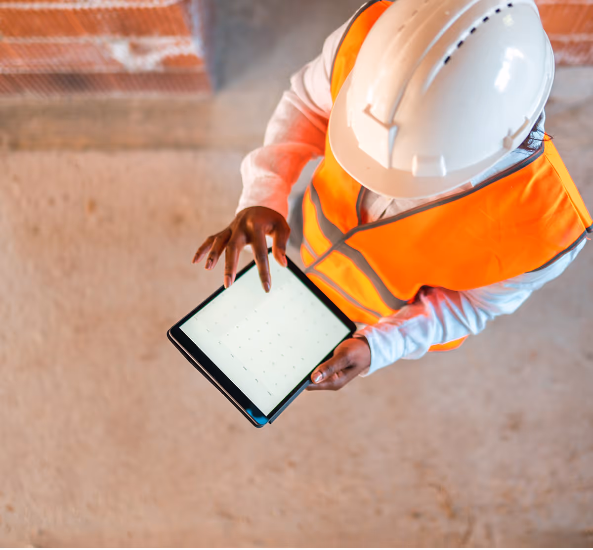 Overhead view of a construction worker in a white hard hat and orange safety vest using a tablet on a job site with a rough concrete floor.