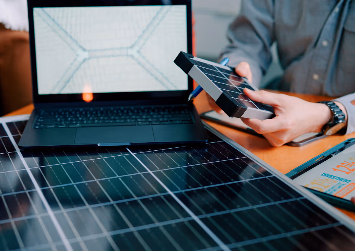 Man at a desk holding a small solar panel while working with a laptop and a larger solar panel, planning or testing solar configurations.
