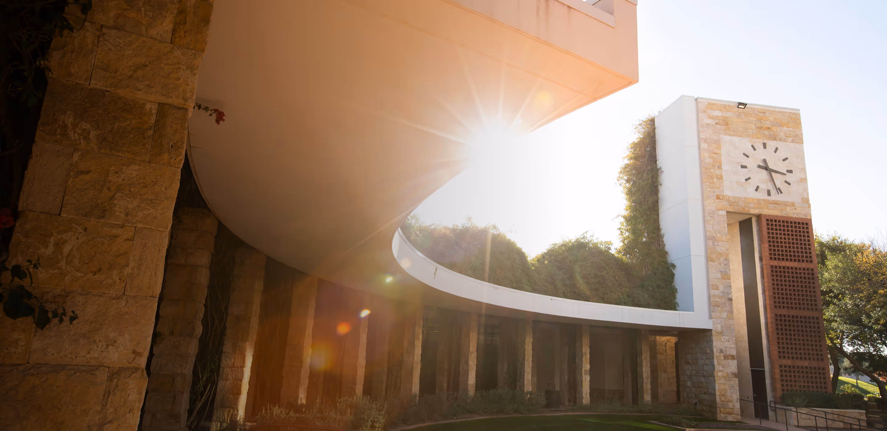 Modern architectural scene with a white clock tower and curving overhang catching sunlight, surrounded by stone columns and greenery.