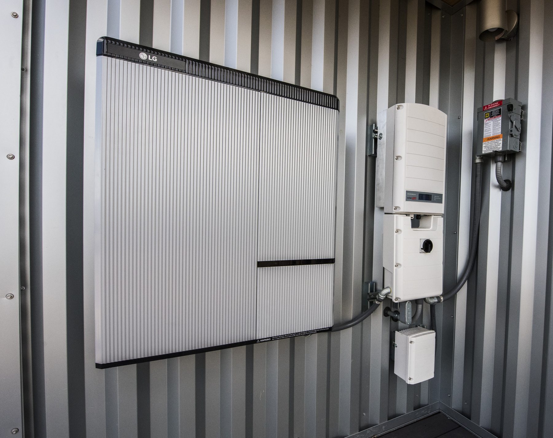A wall-mounted LG home battery installed beside a SolarEdge inverter and PV system disconnect on a corrugated metal wall.