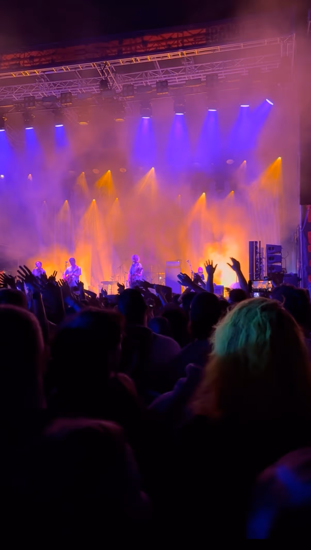 Band performing on a brightly lit stage with orange and purple lights, as a large crowd raises hands and dances in front of them.