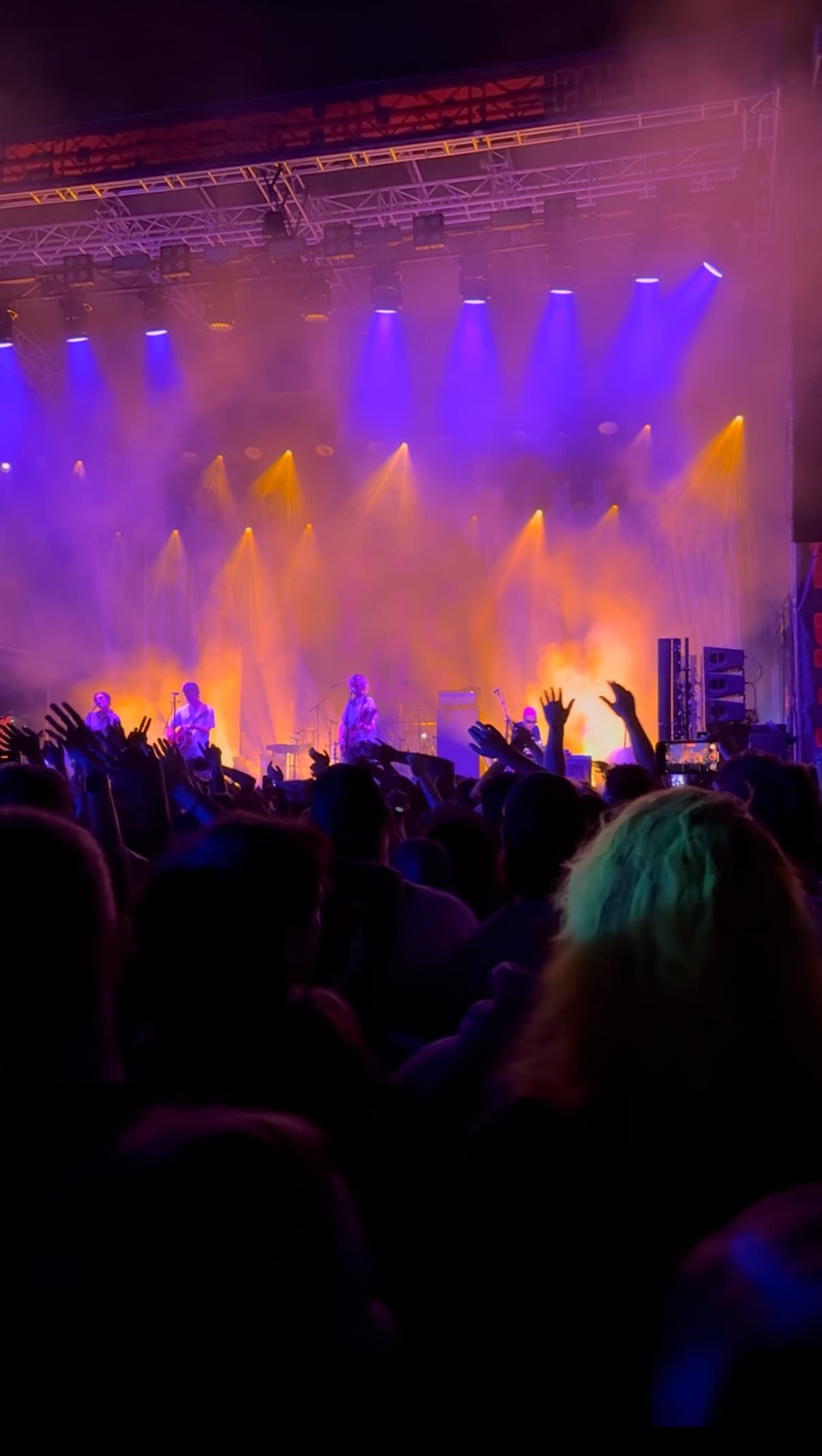 Band performing on a brightly lit stage with orange and purple lights, as a large crowd raises hands and dances in front of them.