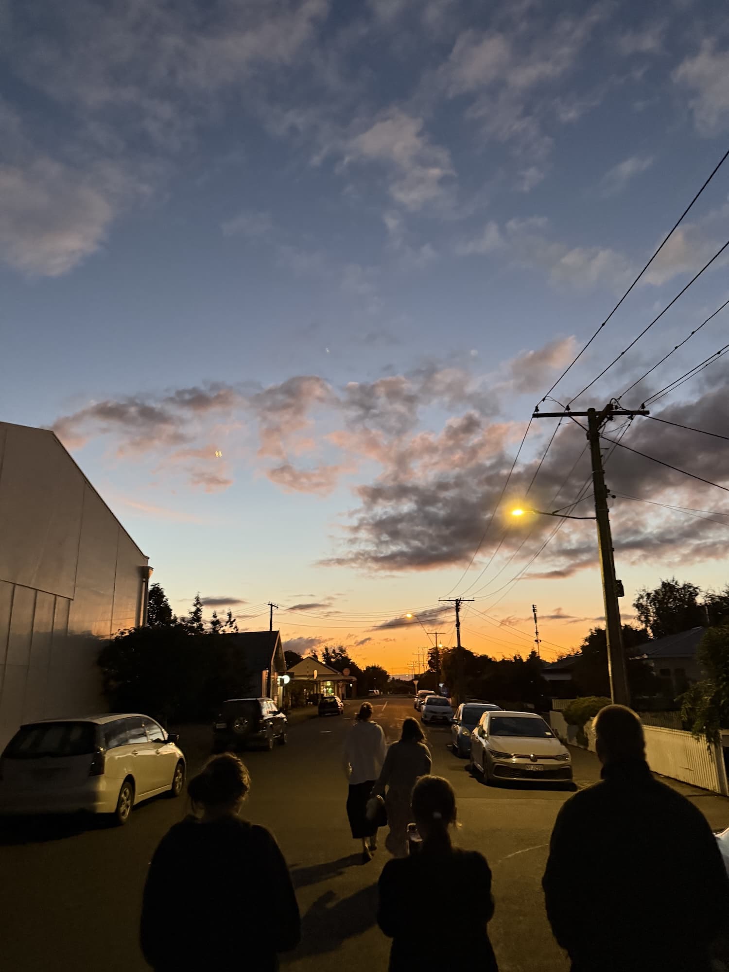 Group of people walking down a quiet suburban street at sunset, with a warm sky, parked cars, and streetlights starting to glow.