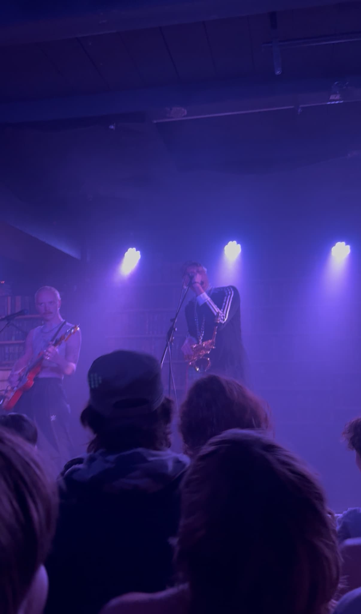 Two musicians performing under purple stage lights in a small venue, one playing saxophone and singing into a mic, the other playing guitar, with an audience watching closely.