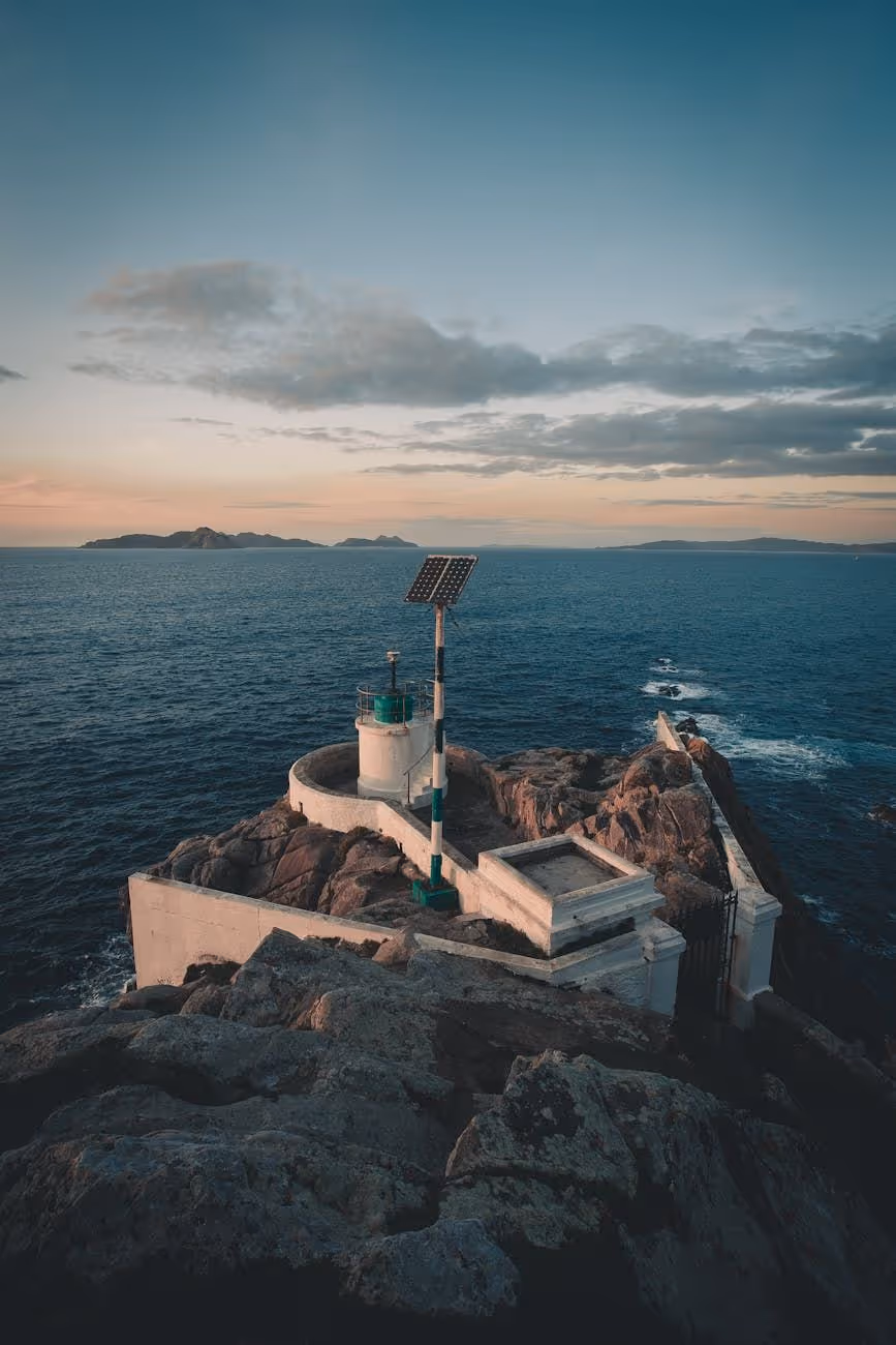 Small lighthouse structure with a solar panel on rocky coastline overlooking calm ocean under a cloudy sky at sunset.