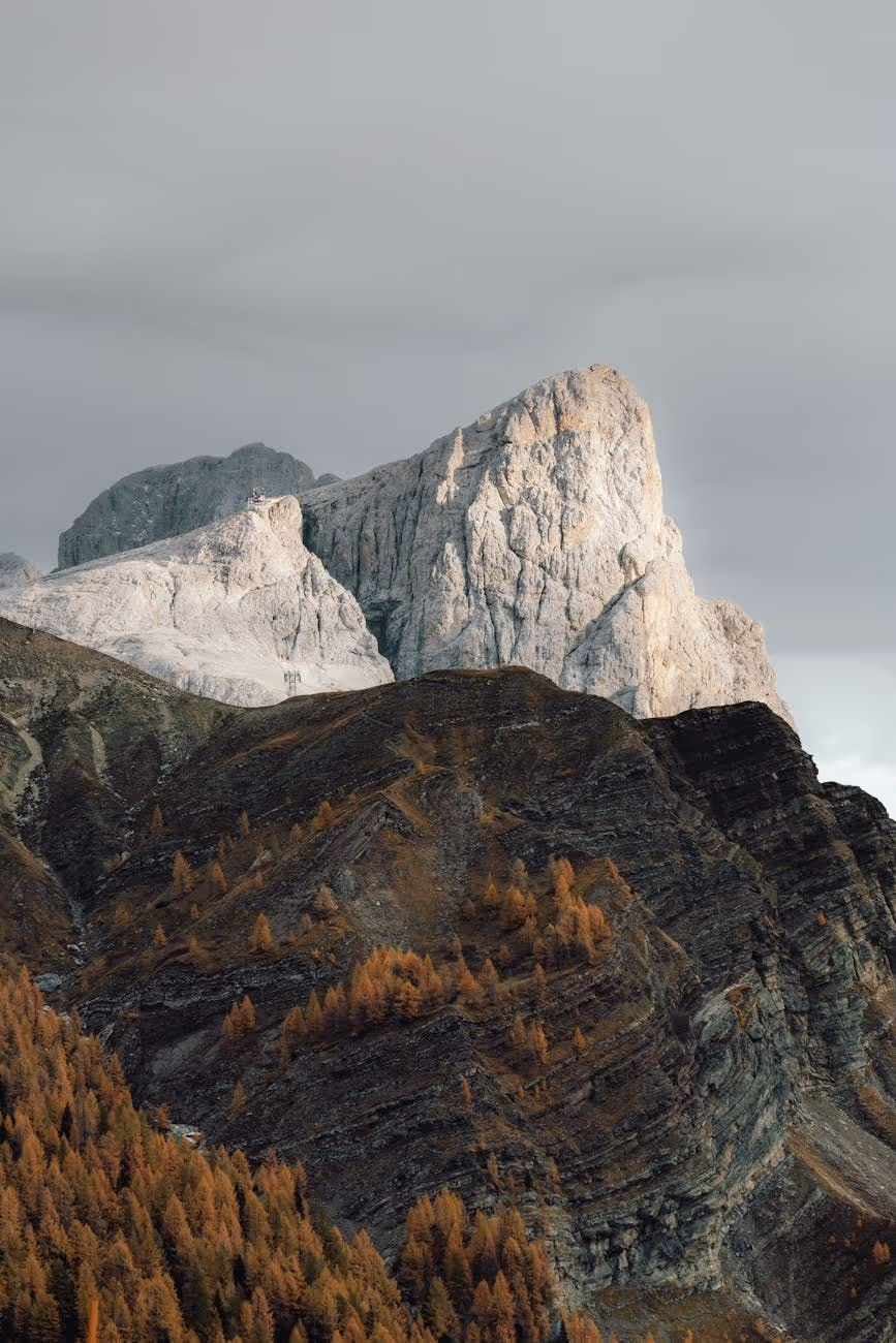 Tall rugged mountain peak illuminated by soft light with dark rocky slopes and autumn-colored trees in the foreground.