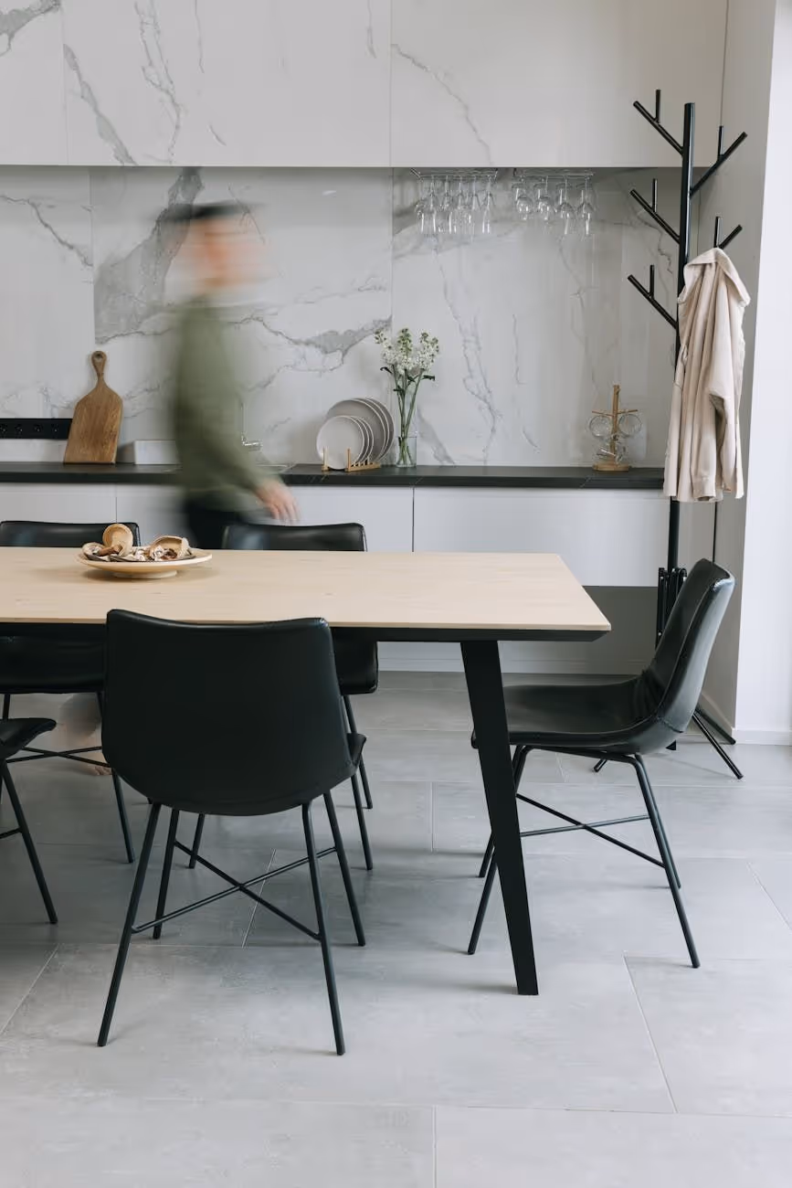 Modern dining area with a light wooden table, black chairs, marble backsplash kitchen counter, and a blurred person walking by.