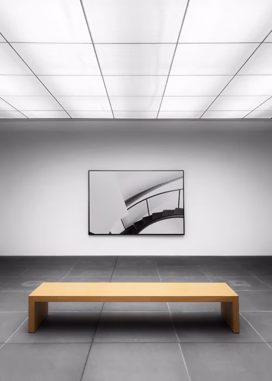 Minimalist gallery room with a wooden bench in front of a black and white photo of a spiral staircase on a white wall.