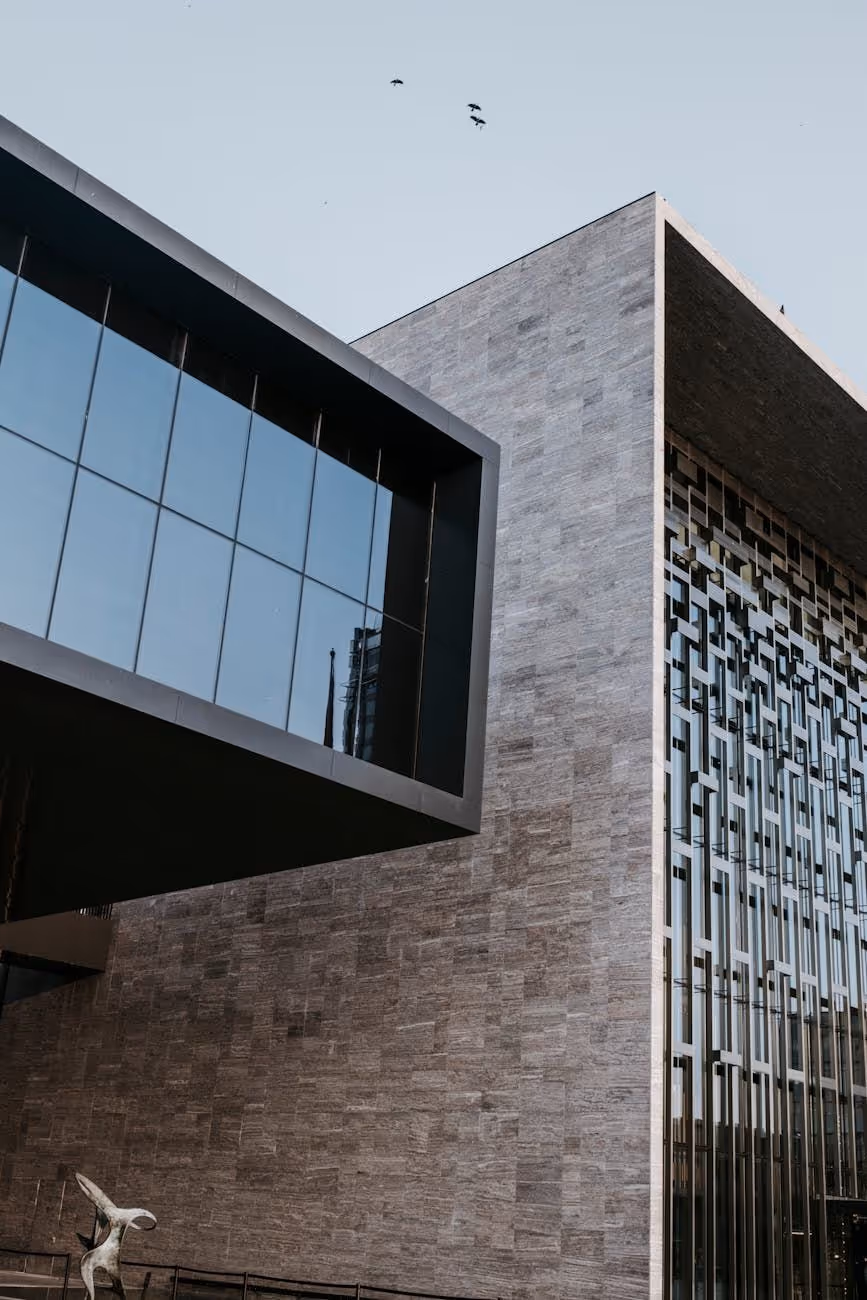 Modern building with large stone facade, reflective glass windows, and a geometric sculpture at the base under a clear sky with flying birds.