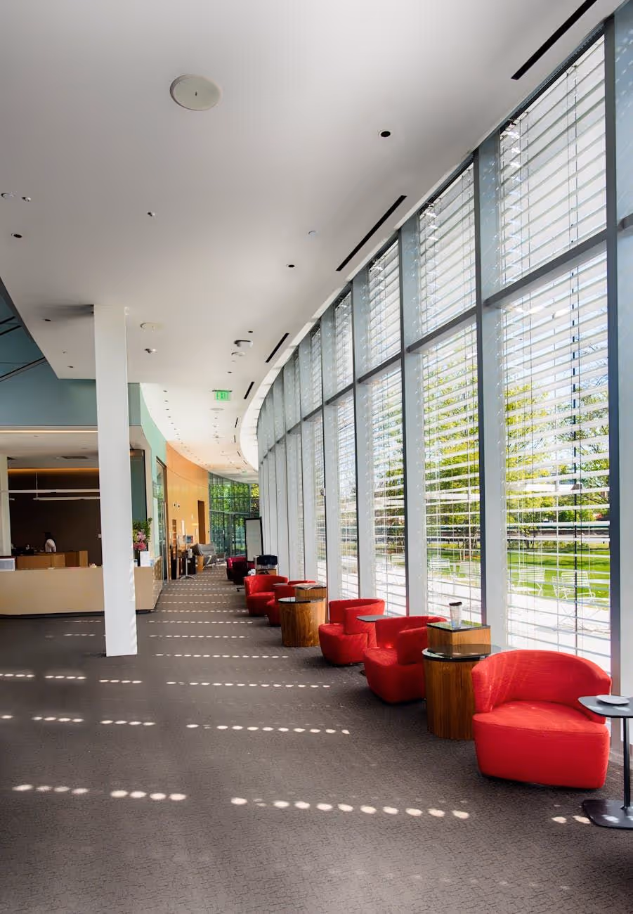 Bright modern lobby with large curved windows, red armchairs, and small wooden tables overlooking a green outdoor space.