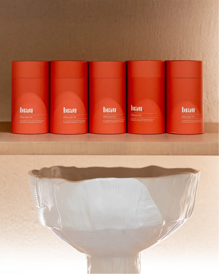 Five orange cylindrical containers labeled 'brau. Aftercare Kit' arranged on a wooden shelf above a white textured ceramic vase.