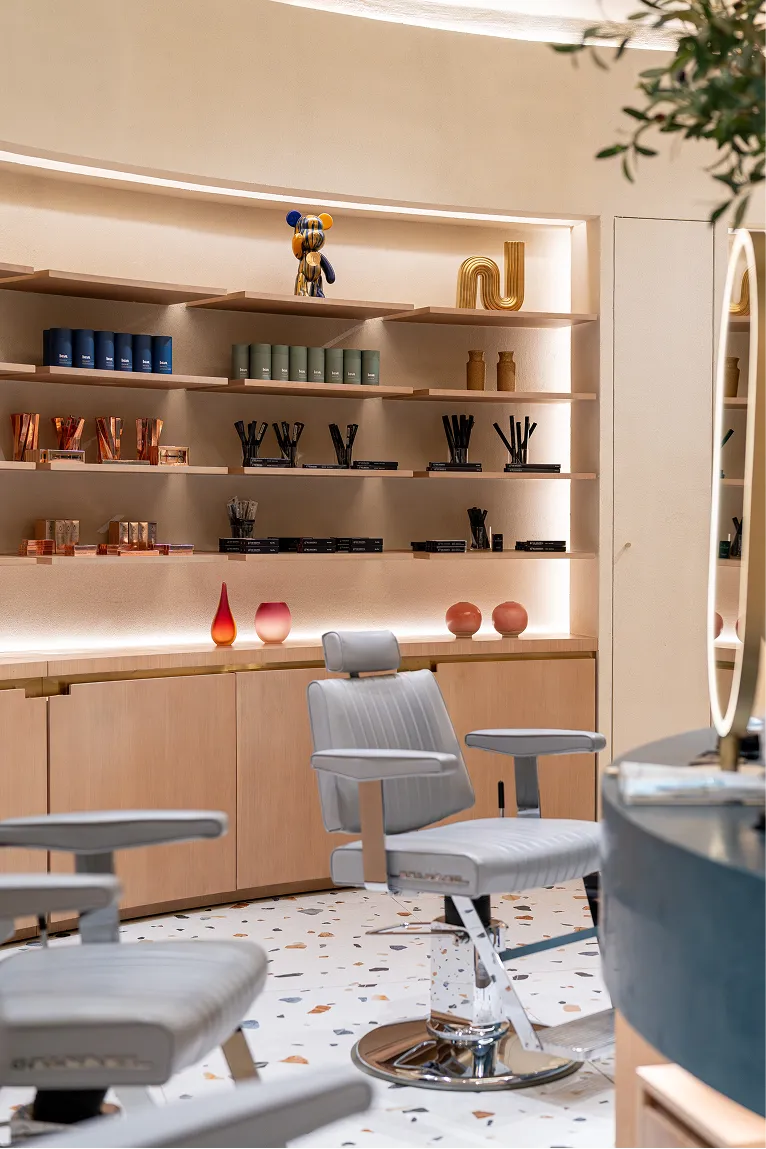 Modern salon interior with white barber chairs, wooden shelves displaying beauty products, decorative vases, and a colorful bear sculpture.