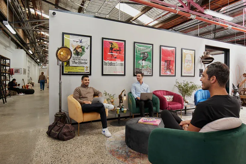 Three men seated in a modern office lounge with laptops, colorful chairs, framed movie posters on the wall, and industrial ceiling.