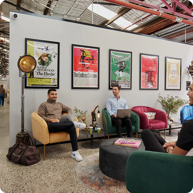 Three men sitting in colorful armchairs in a modern office lounge with laptops, framed movie posters on the wall, and a round coffee table with books.