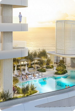 Modern luxury resort with a pool, lounge chairs, greenery, and ocean view at sunset, with a person standing on a balcony.