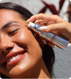 Smiling woman applying serum to her cheek from a silver pump bottle under sunlight.
