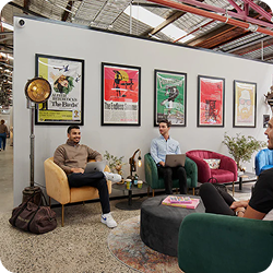 Three men sitting and chatting in a modern lounge area with colorful vintage posters on the wall behind them.