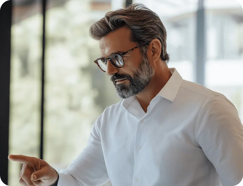 A stylish, middle-aged man with a beard and glasses, wearing a white shirt, points downwards.