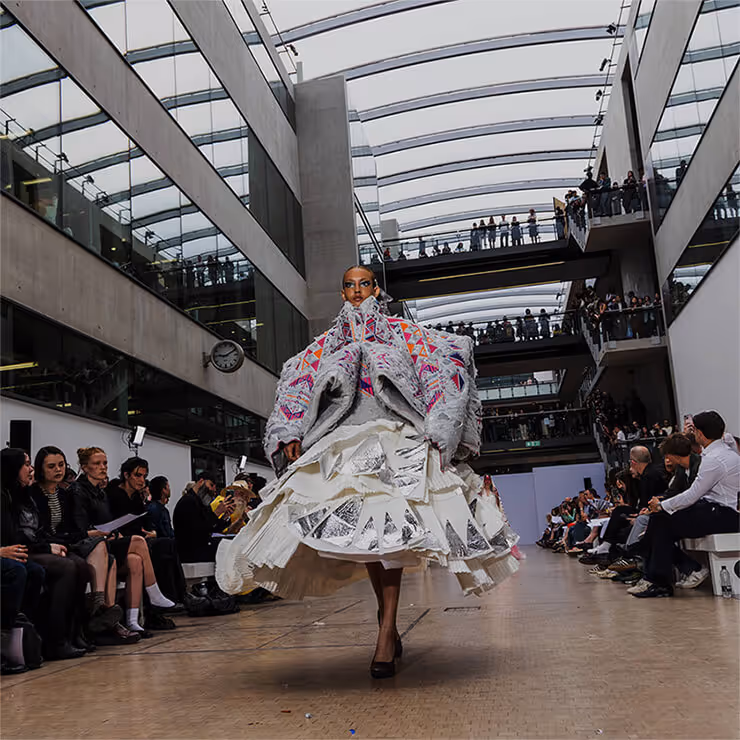 A fashion model walks the catwalk in a couture dress at King's Cross, London. The backdrop features the area's iconic industrial architecture, blending historic brickwork with modern design elements.