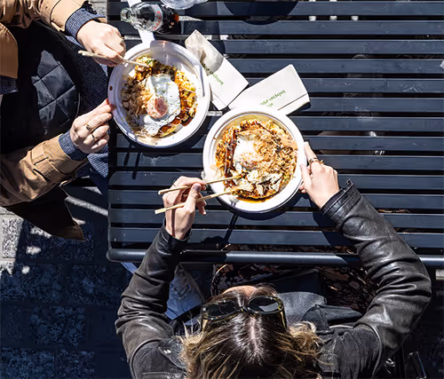 Two people dining outdoors at a black slatted table, eating from bowls with chopsticks. A napkin is visible, along with a bottle of water. Both individuals are wearing jackets and sunglasses, suggesting a casual urban setting.