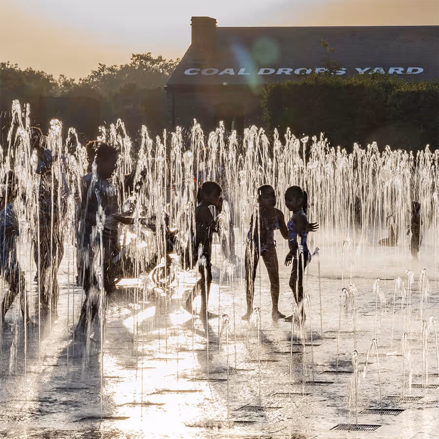 Children joyfully run through and play in the fountains at Granary Square, King's Cross, on a bright summer day, surrounded by urban architecture and open public space.
