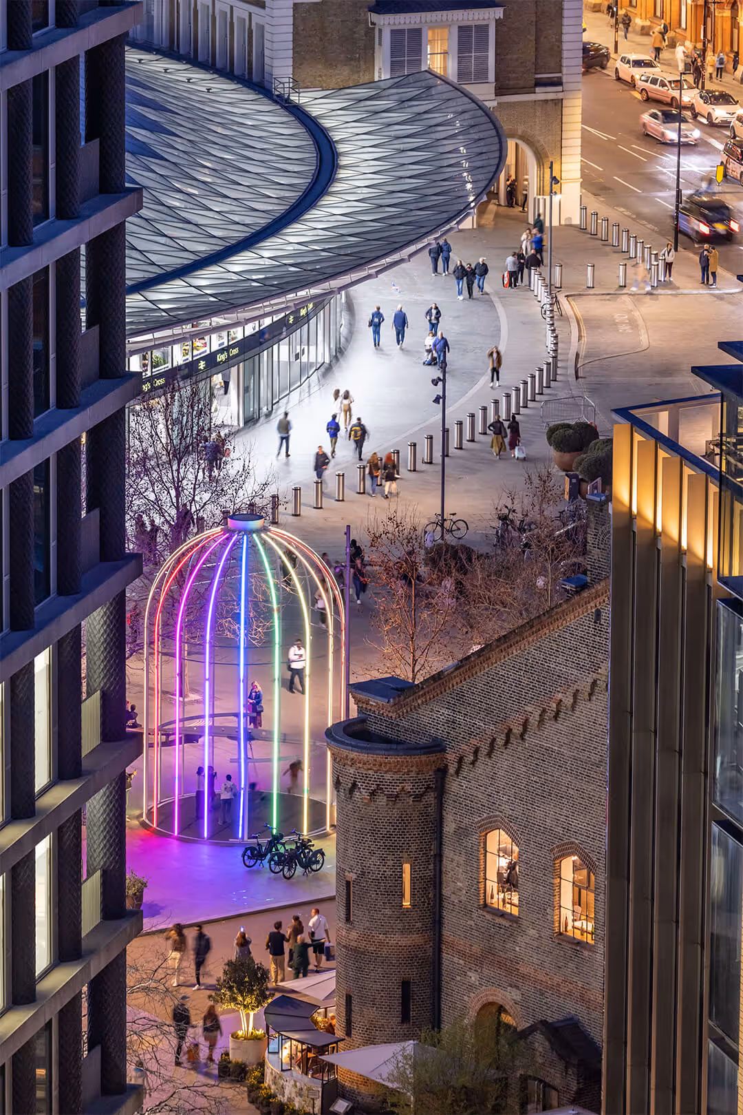 It's night time. A high-up shot of the rainbow-lit swing that sits outside Kings Cross station illuminates the area.