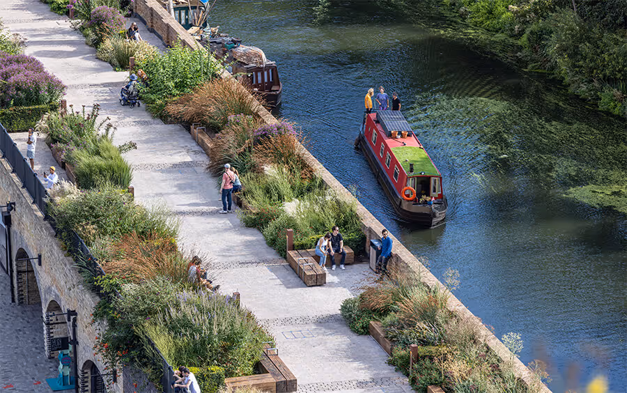 A canal boat gently drifts through the clear canal water as small groups of people walk relaxingly across the banks in the Coal Drop Yard area of King's Cross.