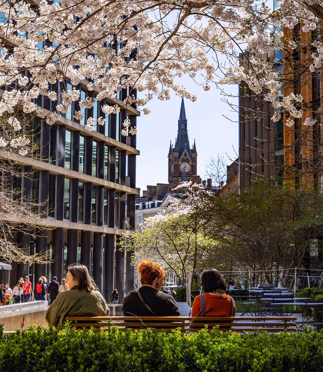Three people sit on a bench on a crisp spring day as they admire the view between the buildings from St Pancras square. The clocktower of St Pancras hotel is large in the background.