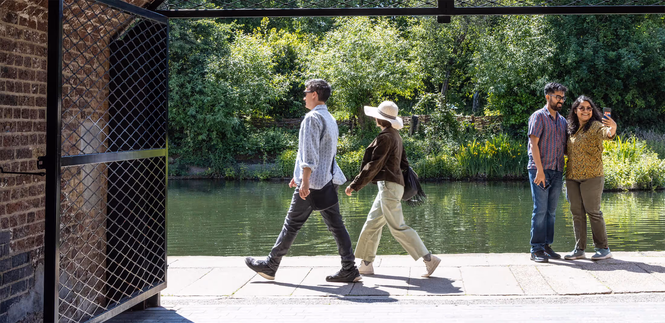 A couple pose for a selfie at the edge of King's Cross canal. Another couple walk hand in hand as they enjoy the warm summers day.