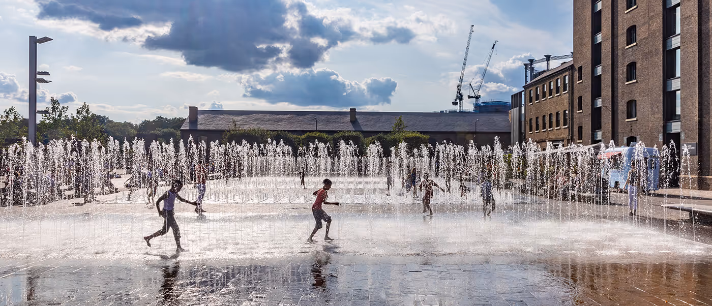 Children happily run through and play in the fountains at King's Cross Granary Square on a sunny summer day.