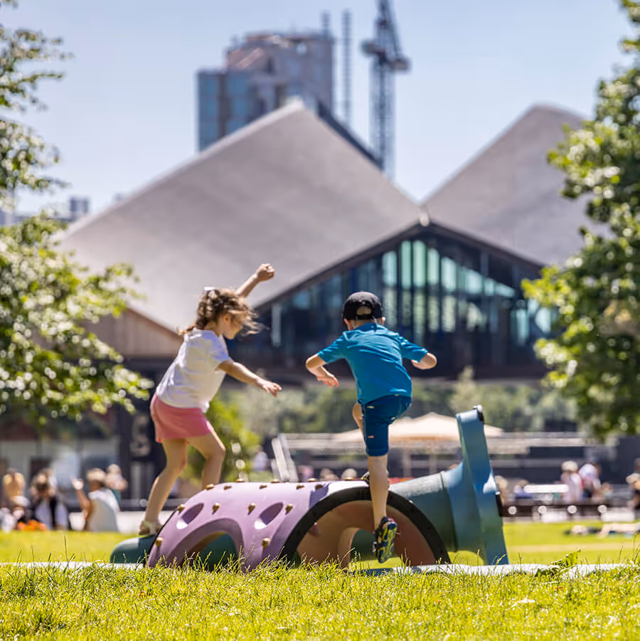Children playing on colourful playground equipment in a sunny park, with the modern Coal Drops Yard building and trees in the background. Other park visitors are visible in the distance.