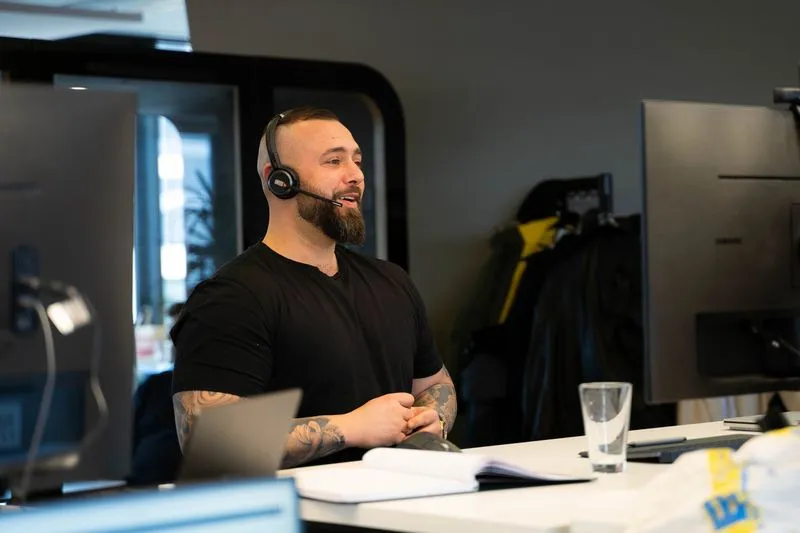 Mann mit Headset sitzt am Schreibtisch, spricht und schaut auf einen Monitor im Büro
