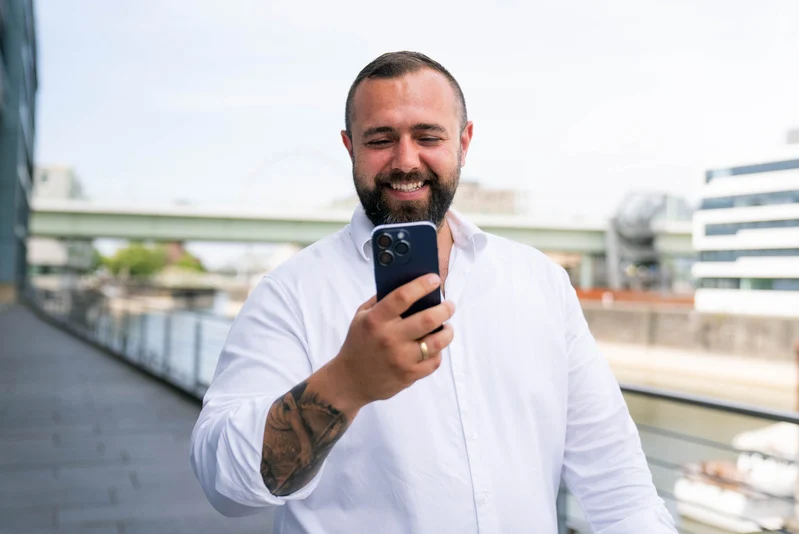 Jan Truch, Geschäftsführer der Headfound Group, hält ein blaues iPhone 15 Pro in der Hand, das im Fokus steht, während er dahinter auf das Handy schaut und lächelt. Das Foto wurde am Rheinauhafen in Köln aufgenommen