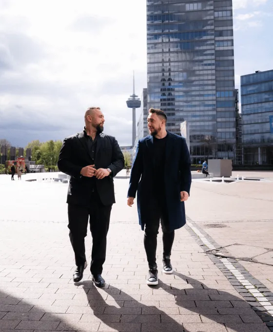 Zwei Männer gehen nebeneinander auf einem Platz vor modernen Bürogebäuden bei sonnigem Wetter
