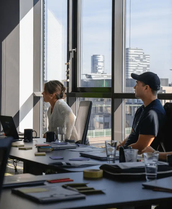 Zwei Personen sitzen in einem modernen Büro am Besprechungstisch und schauen konzentriert in Richtung eines Bildschirms mit Stadtblick im Hintergrund