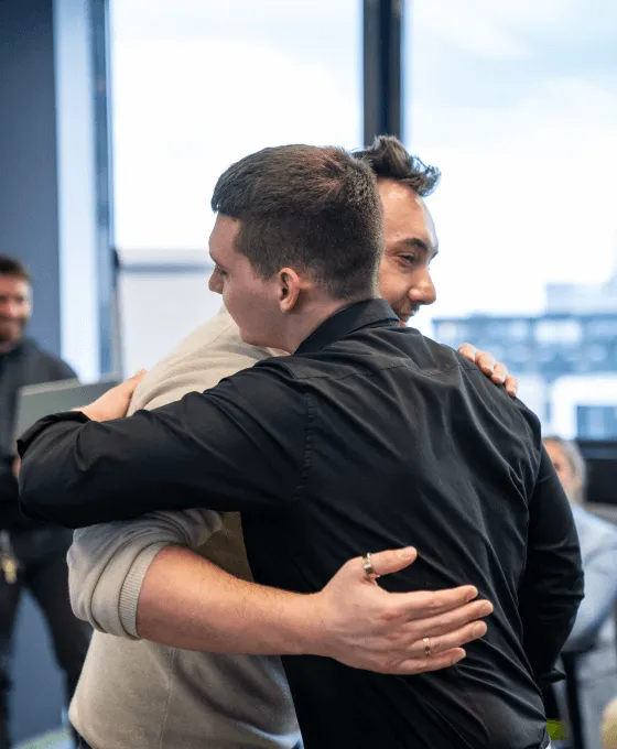 Zwei Männer umarmen sich herzlich in einem modernen Büro mit Fensterfront im Hintergrund