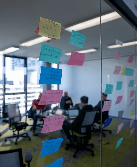 Besprechungsraum mit Glaswand voller bunter Notizzettel im Vordergrund und Teammeeting im Hintergrund