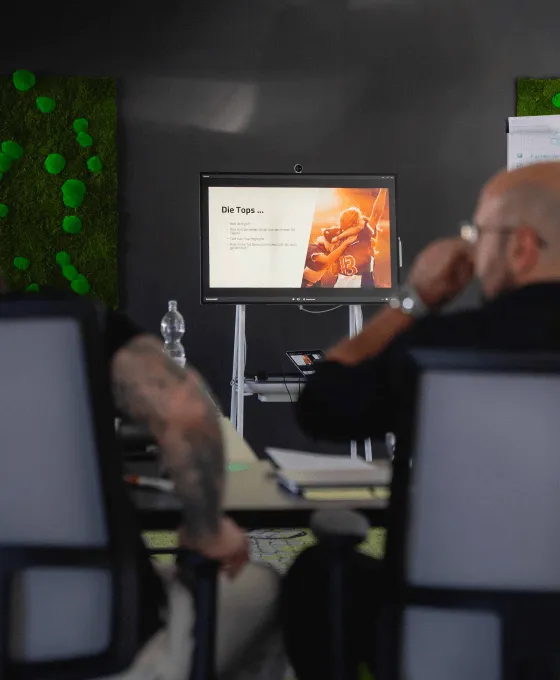 Zwei Männer sitzen in einem Besprechungsraum vor einem Bildschirm, auf dem eine Präsentation angezeigt wird