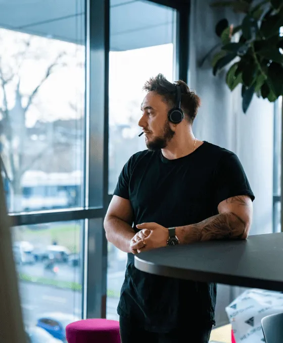 Mann mit Headset steht an einem Stehtisch vor großen Fenstern und schaut nach draußen