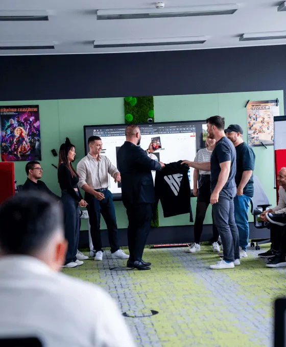 Mehrere Personen stehen in einem modernen Raum, während ein Mann ein schwarzes T-Shirt mit Logo übergibt