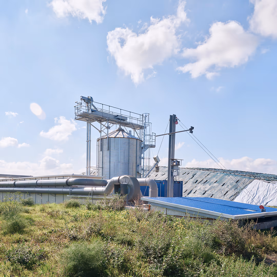 Industrielle Anlage mit Metallbehälter, Rohrleitungen und Solarpanelen vor bewachsenem Boden unter blauem Himmel mit Wolken.