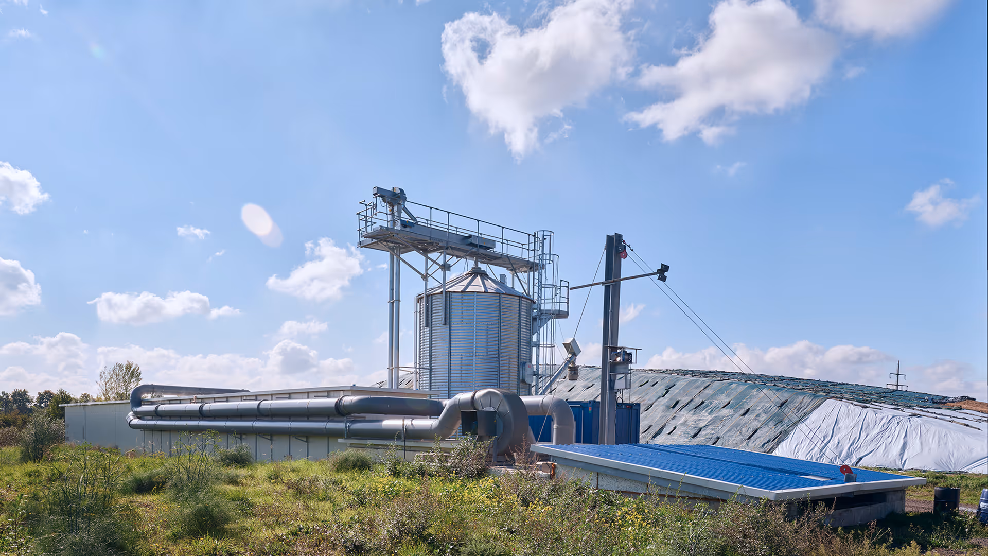 Industrielle Anlage mit großem Metallbehälter, Rohrleitungen und mit Kunststoffplanen bedecktem Berg unter blauem Himmel mit vereinzelten Wolken.