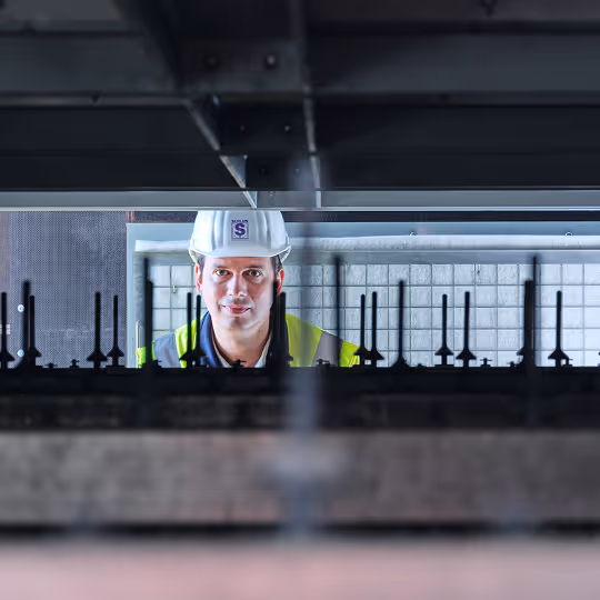 Bauarbeiter mit Schutzhelm und Warnweste blickt durch Metallstruktur auf einer Baustelle.
