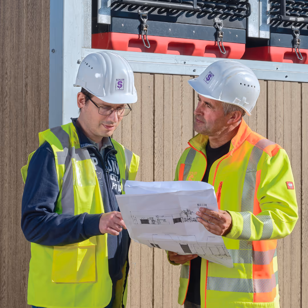 Zwei Bauarbeiter mit weißen Helmen und neonfarbenen Warnwesten, die Baupläne gemeinsam betrachten.