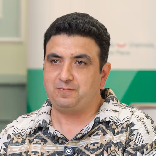 Man with short curly black hair wearing a patterned button-up shirt against a blurred indoor background.