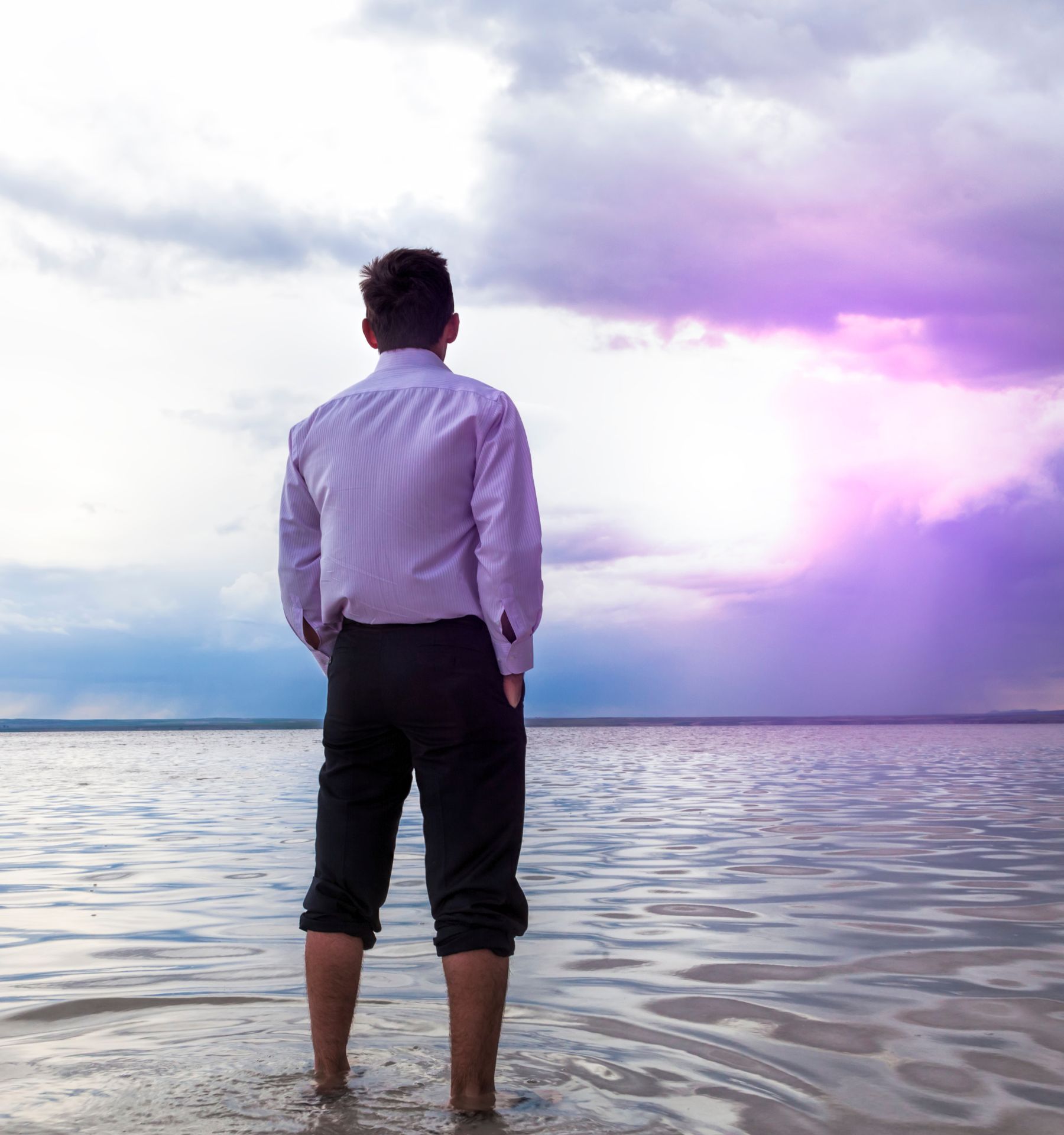 A man is standing in the water with his trousers rolled up to his knees looking toward the horizon