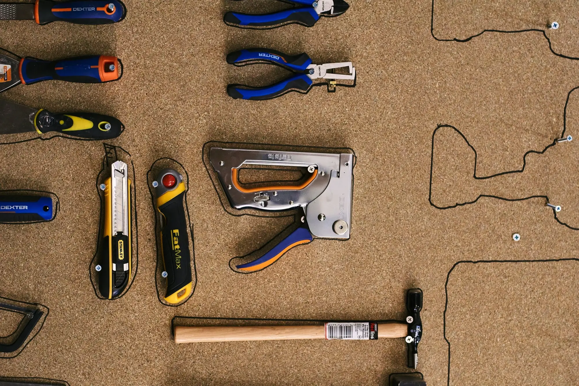 Outils à main organisés sur un panneau en liège, incluant un marteau, des pinces, un cutter et une agrafeuse, fixés et entourés par des contours noirs.