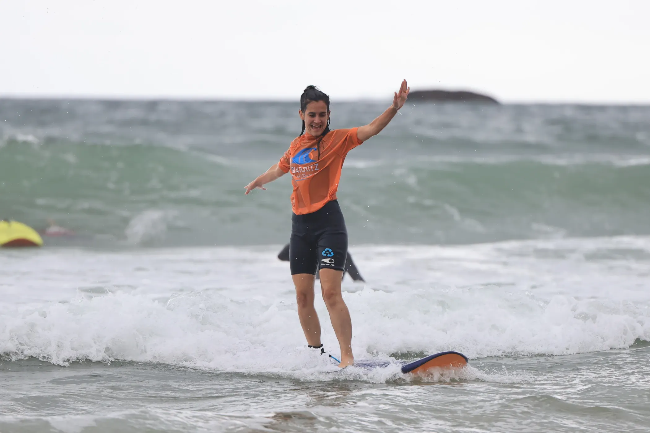 Femme souriante en maillot de bain et T-shirt orange faisant du surf sur une planche courte dans une mer agitée.