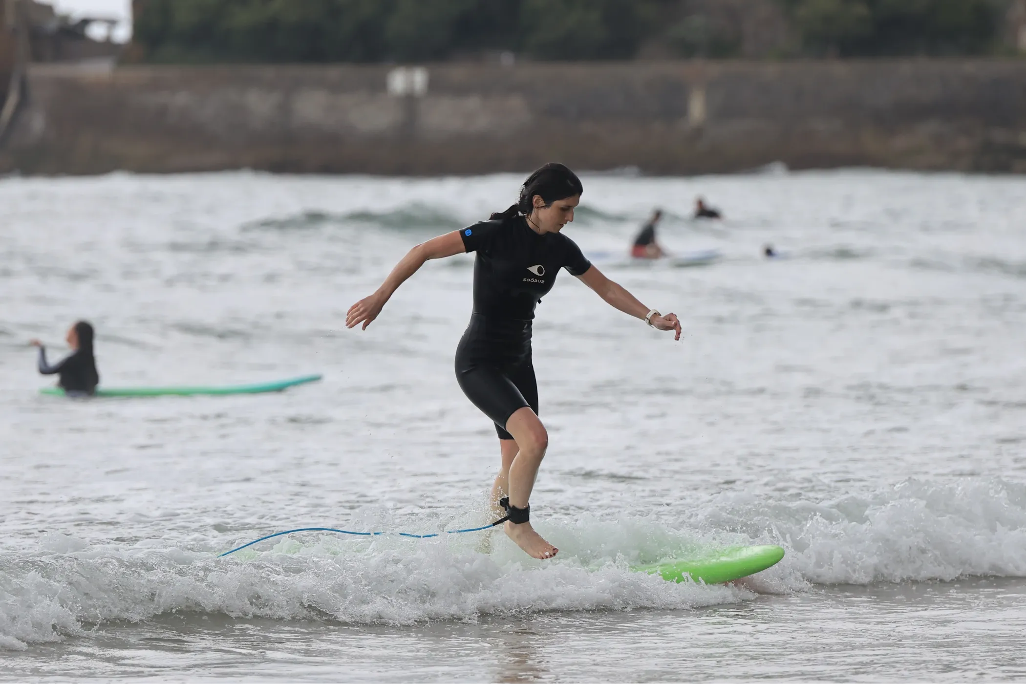 Une femme en combinaison de surf noire équilibre sur une planche verte dans les vagues peu profondes de l'océan.